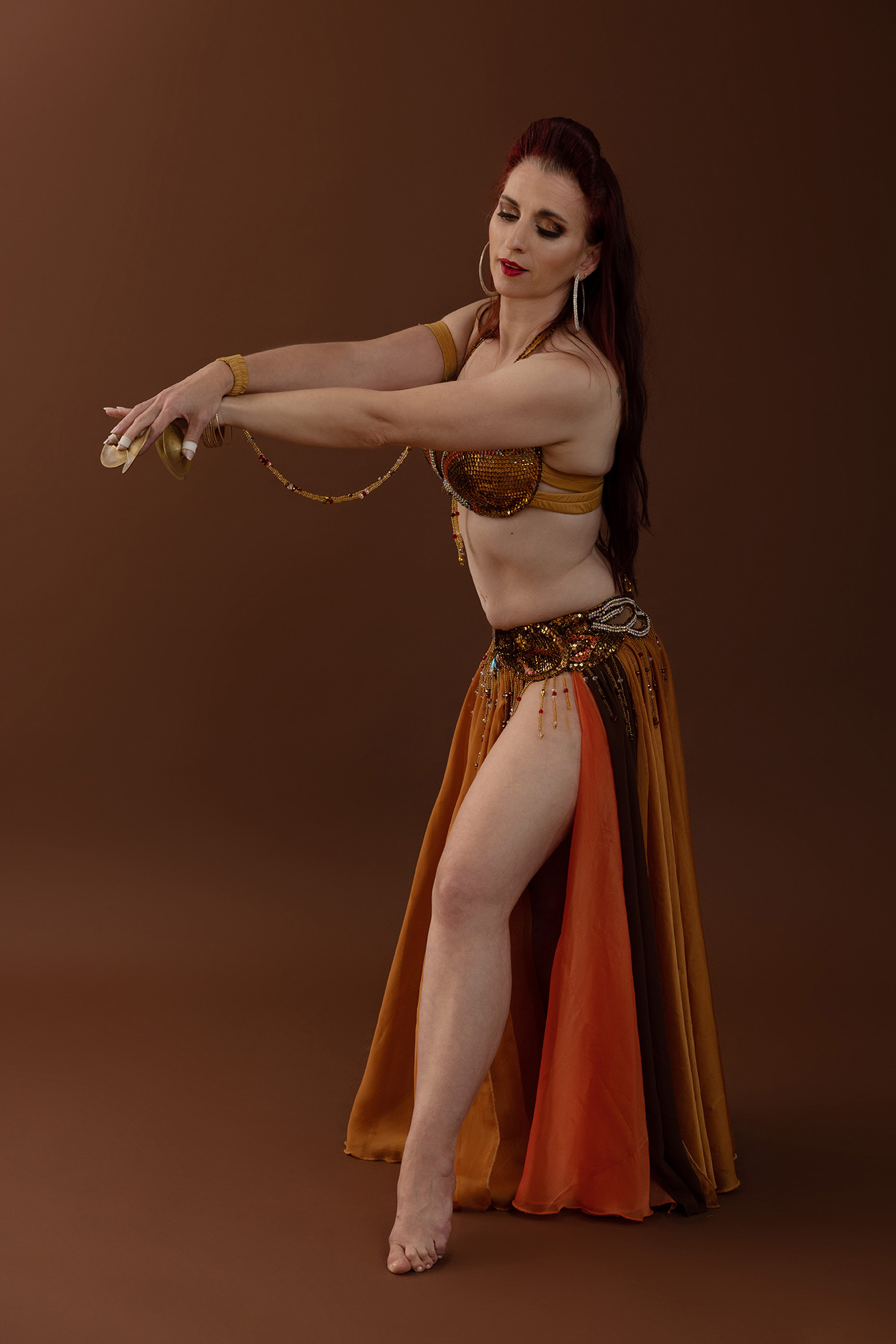 Amanda posting in an orange 2 piece belly dance costume, facing to the left with both arms extended towards the left with finger cymbals on her fingers. She has one leg out from a split in the skirt.